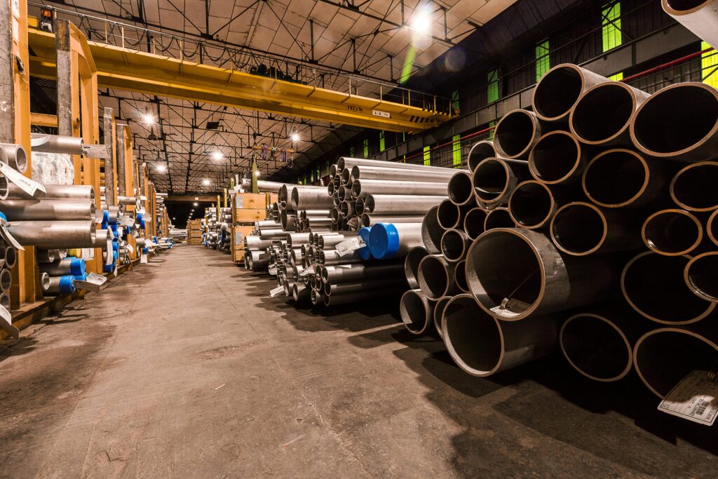 Interior of an industrial warehouse with stacks of metal pipes and high ceilings.