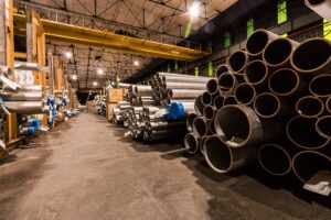 Interior of an industrial warehouse with stacks of metal pipes and high ceilings.